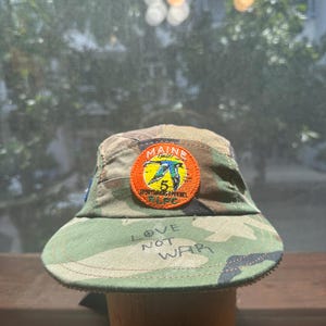 May include: A camouflage print five-panel cap featuring an orange circular patch with a blue bird illustration, the text MAINE, 5, SPORTSMAN APPAREL, and RLPC. The brim is embroidered with the text LOVE NOT WAR in a dark green thread.