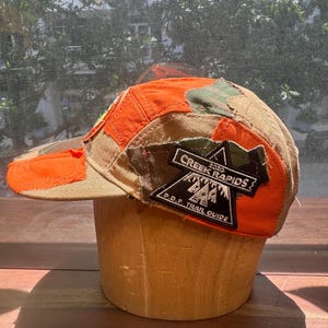 Peut inclure: Une casquette &agrave; cinq panneaux avec un design en patchwork de tissu orange vif et camouflage. Un &eacute;cusson triangulaire noir sur le c&ocirc;t&eacute; affiche le texte blanc 2020 CREEK RAPIDS P.D.F. TRAIL GUIDE avec un graphique de montagnes et de sapins.