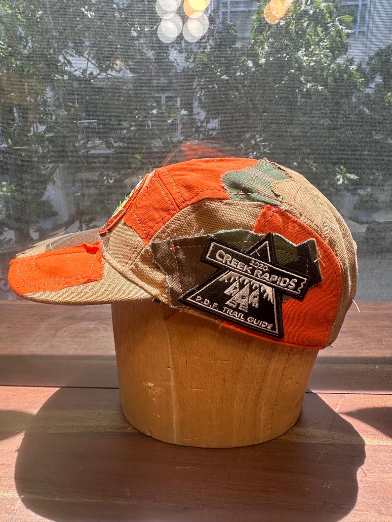 Peut inclure: Une casquette &agrave; cinq panneaux avec un design en patchwork de tissu orange vif et camouflage. Un &eacute;cusson triangulaire noir sur le c&ocirc;t&eacute; affiche le texte blanc 2020 CREEK RAPIDS P.D.F. TRAIL GUIDE avec un graphique de montagnes et de sapins.
