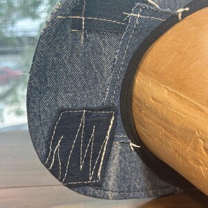 May include: A blue denim hat featuring a patchwork design with visible white stitching. The hat is constructed from multiple shades of denim fabric, including a darker blue rectangular patch, and is finished with a black inner band lining.
