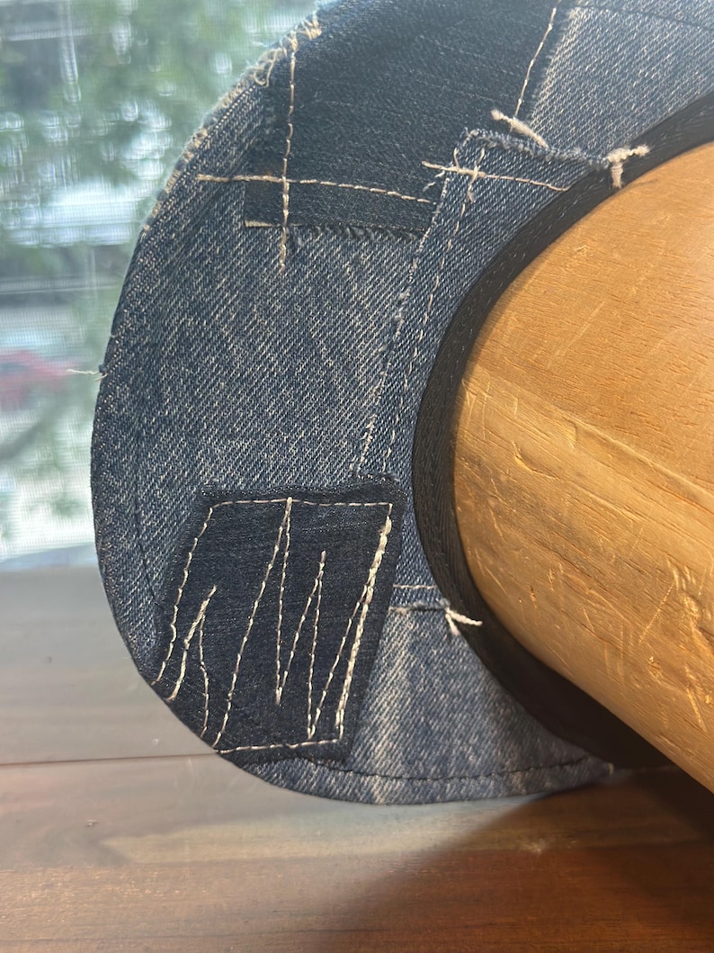 May include: A blue denim hat featuring a patchwork design with visible white stitching. The hat is constructed from multiple shades of denim fabric, including a darker blue rectangular patch, and is finished with a black inner band lining.