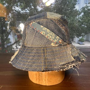 May include: A patchwork bucket hat constructed from various shades of blue denim fabric. The hat features visible frayed edges, yellow topstitching, and a rectangular fabric patch with a light blue floral pattern attached with red stitching.