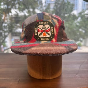 May include: A five-panel cap featuring a multi-colored patterned fabric in shades of brown, red, and blue. A central patch displays the text FOREST RIVER CAMP GUIDE with an embroidered graphic of a pine tree, crossed oars, and a campfire.
