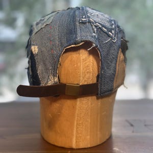May include: A baseball cap constructed from various shades of blue denim fabric with visible frayed edges and decorative white stitching. The cap features a brown leather adjustable strap at the back secured with a rectangular brass-toned metal buckle.