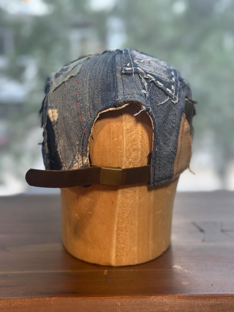 May include: A baseball cap constructed from various shades of blue denim fabric with visible frayed edges and decorative white stitching. The cap features a brown leather adjustable strap at the back secured with a rectangular brass-toned metal buckle.