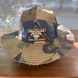 K&ouml;nnte beinhalten: Ein mehrteiliger Fischerhut mit einem Patchwork-Design aus Camouflage- und blauem Jeansstoff. Ein schwarzer dreieckiger Aufn&auml;her ist vorne mittig angebracht und zeigt den wei&szlig;en Text 2020 CREEK RAPIDS und P.D.F. TRAIL GUIDE mit einem Bergsymbol.