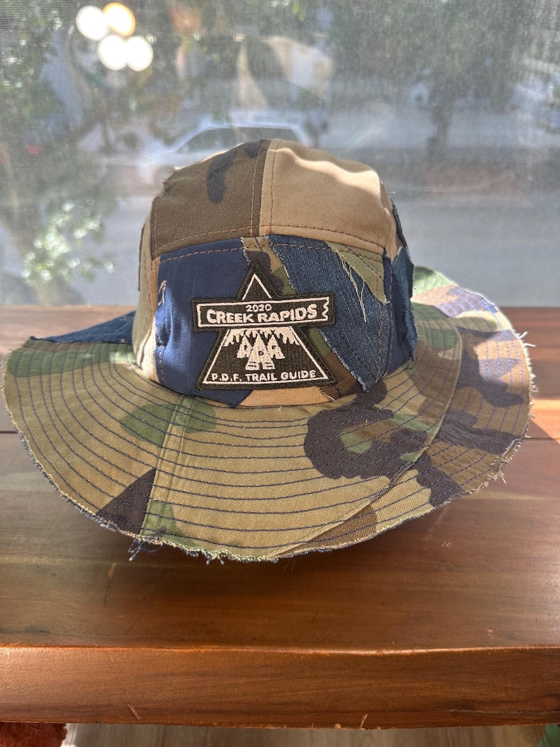 K&ouml;nnte beinhalten: Ein mehrteiliger Fischerhut mit einem Patchwork-Design aus Camouflage- und blauem Jeansstoff. Ein schwarzer dreieckiger Aufn&auml;her ist vorne mittig angebracht und zeigt den wei&szlig;en Text 2020 CREEK RAPIDS und P.D.F. TRAIL GUIDE mit einem Bergsymbol.