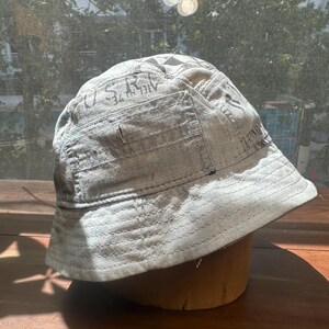 May include: A light grey bucket hat featuring a distressed, weathered texture with visible stitched seams and dark printed lettering that includes the text U.S.R. and other partial graphics across the crown and side panels of the fabric.