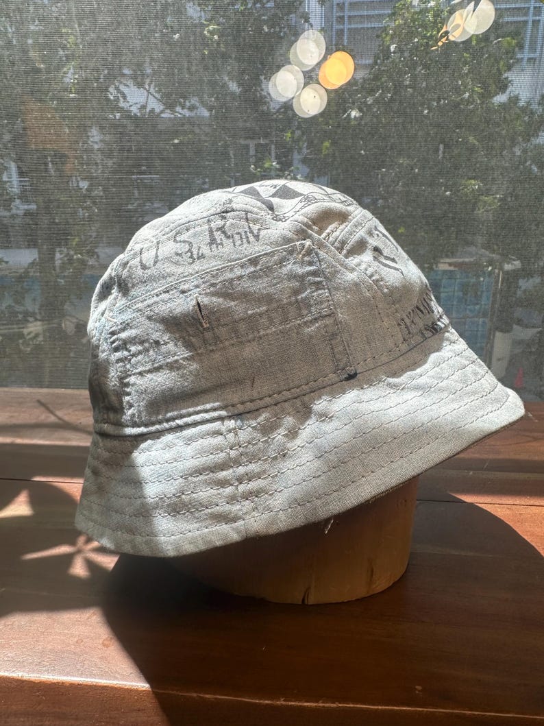 May include: A light grey bucket hat featuring a distressed, weathered texture with visible stitched seams and dark printed lettering that includes the text U.S.R. and other partial graphics across the crown and side panels of the fabric.