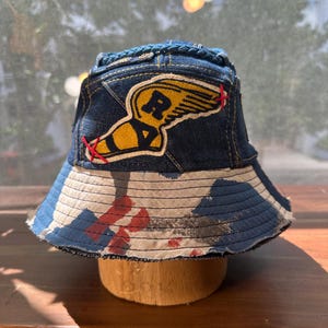 May include: A bucket hat constructed from dark blue denim and a light-coloured fabric with a blue and red painted pattern. The crown features a yellow winged shoe patch with the letter R in black, accented by red cross-stitch details on the sides.