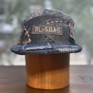 May include: A five-panel cap constructed from dark blue denim patchwork fabric with visible light-colored stitching. A rectangular fabric patch on the front features the text RL,-5.8.A-5 in a light-colored font against a dark background.