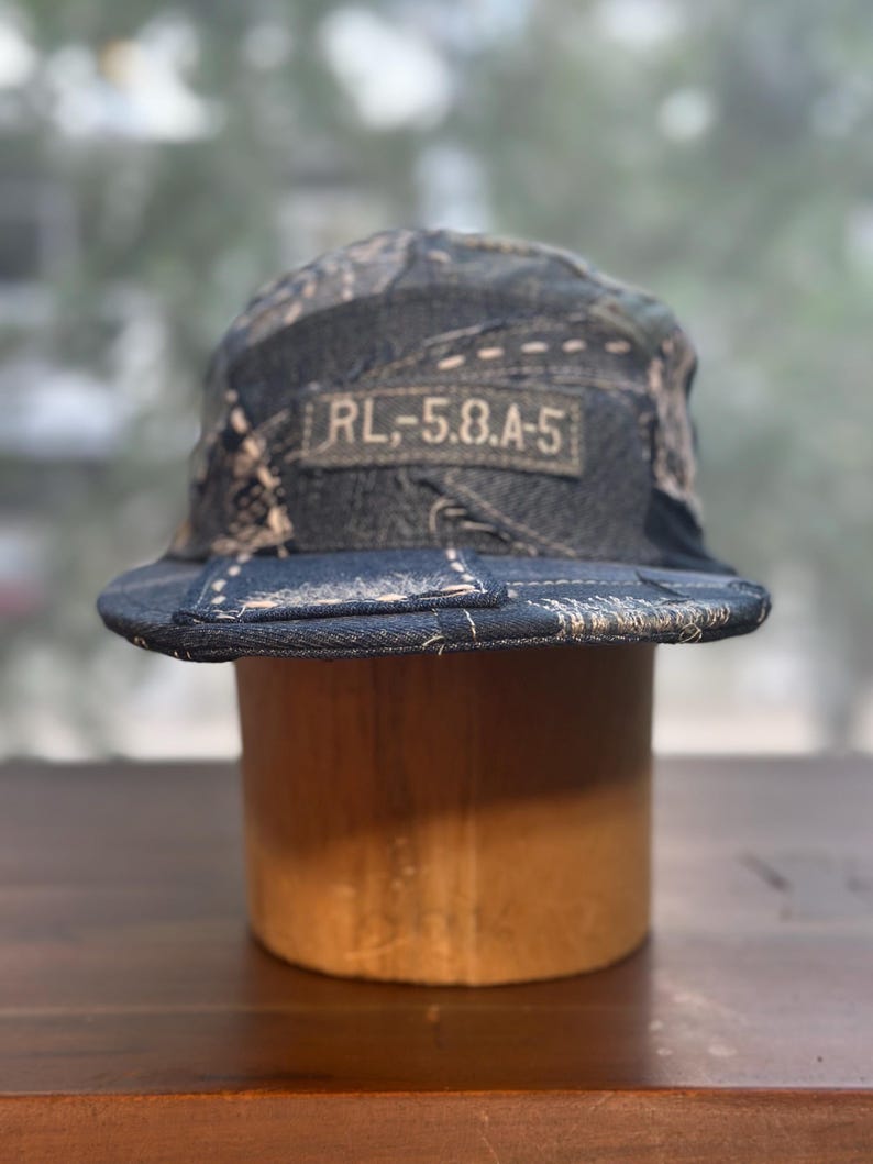 May include: A five-panel cap constructed from dark blue denim patchwork fabric with visible light-colored stitching. A rectangular fabric patch on the front features the text RL,-5.8.A-5 in a light-colored font against a dark background.