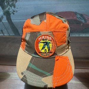 Peut inclure: Une casquette de baseball au design patchwork compos&eacute; de tissu orange vif et camouflage. Un &eacute;cusson brod&eacute; circulaire sur le devant affiche une silhouette de canard vert, un soleil jaune et le texte MAINE, 5, SPORTSMANS APPAREL et RLPC.