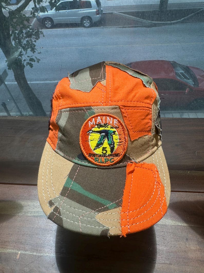 Peut inclure: Une casquette de baseball au design patchwork compos&eacute; de tissu orange vif et camouflage. Un &eacute;cusson brod&eacute; circulaire sur le devant affiche une silhouette de canard vert, un soleil jaune et le texte MAINE, 5, SPORTSMANS APPAREL et RLPC.