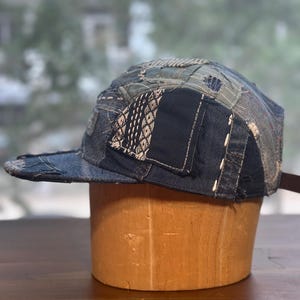 May include: A five-panel cap constructed from various shades of blue denim fabric. The hat features a patchwork design with visible white sashiko-style stitching, frayed edges, and multiple fabric textures layered across the crown and brim.