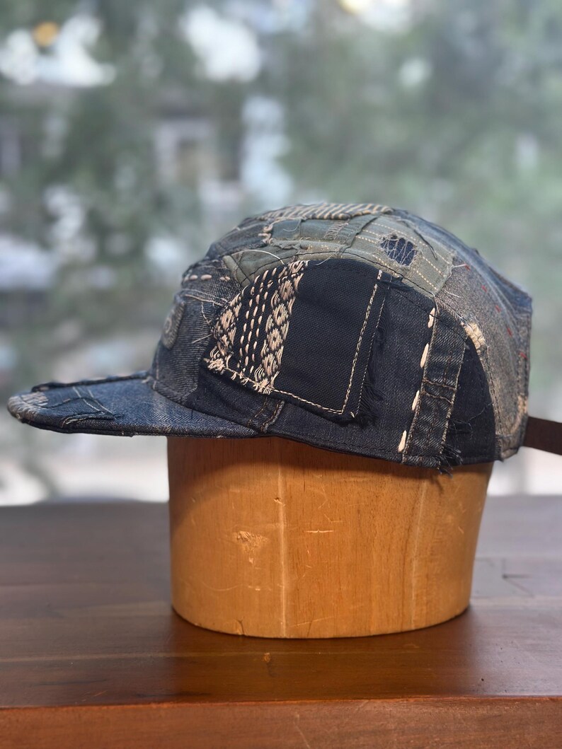 May include: A five-panel cap constructed from various shades of blue denim fabric. The hat features a patchwork design with visible white sashiko-style stitching, frayed edges, and multiple fabric textures layered across the crown and brim.
