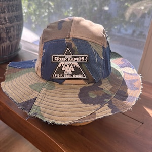 K&ouml;nnte beinhalten: Ein Patchwork-Anglerhut mit Camouflage-Muster in Gr&uuml;n-, Braun- und Blaut&ouml;nen. Auf der Vorderseite befindet sich ein schwarzer, dreieckiger Aufn&auml;her mit wei&szlig;em Text 2020 CREEK RAPIDS und P.D.F. TRAIL GUIDE &uuml;ber einer Berggrafik.