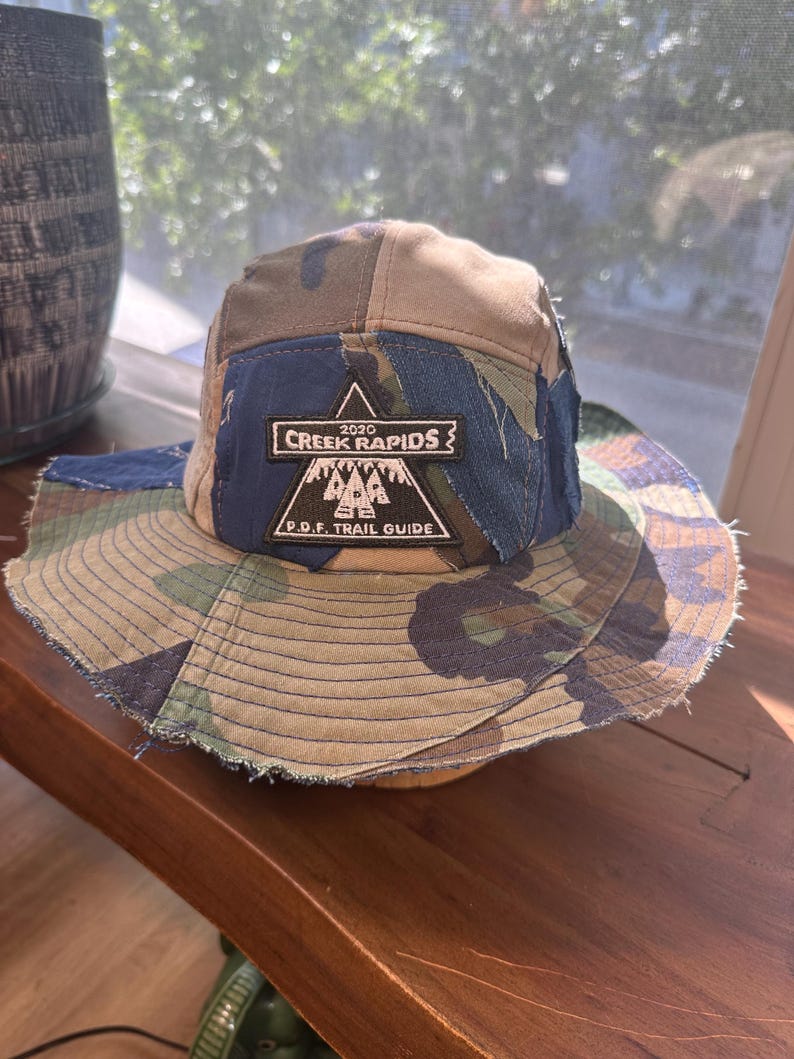 K&ouml;nnte beinhalten: Ein Patchwork-Anglerhut mit Camouflage-Muster in Gr&uuml;n-, Braun- und Blaut&ouml;nen. Auf der Vorderseite befindet sich ein schwarzer, dreieckiger Aufn&auml;her mit wei&szlig;em Text 2020 CREEK RAPIDS und P.D.F. TRAIL GUIDE &uuml;ber einer Berggrafik.