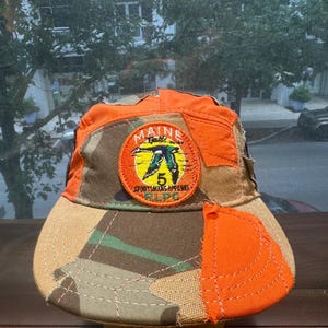 Peut inclure: Une casquette de baseball avec un motif patchwork en tissu orange vif et camouflage. Un &eacute;cusson brod&eacute; circulaire sur le devant affiche une illustration de canard, le texte MAINE, le chiffre 5, SPORTSMANS APPAREL et RLPC en orange.