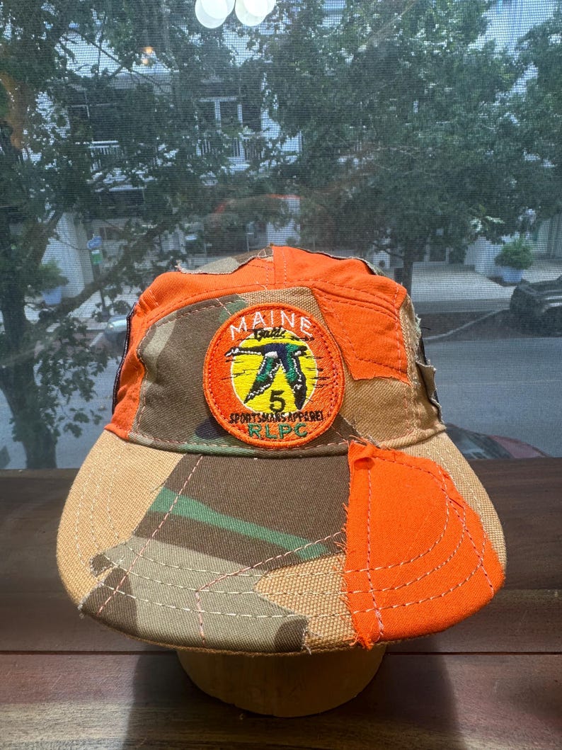 Peut inclure: Une casquette de baseball avec un motif patchwork en tissu orange vif et camouflage. Un &eacute;cusson brod&eacute; circulaire sur le devant affiche une illustration de canard, le texte MAINE, le chiffre 5, SPORTSMANS APPAREL et RLPC en orange.