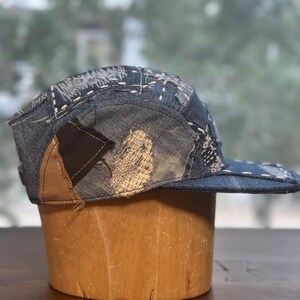 May include: A five-panel cap constructed from various shades of blue denim fabric. The hat features visible white sashiko-style stitching, frayed edges, and contrasting brown and black fabric patches integrated into the side and back panels.