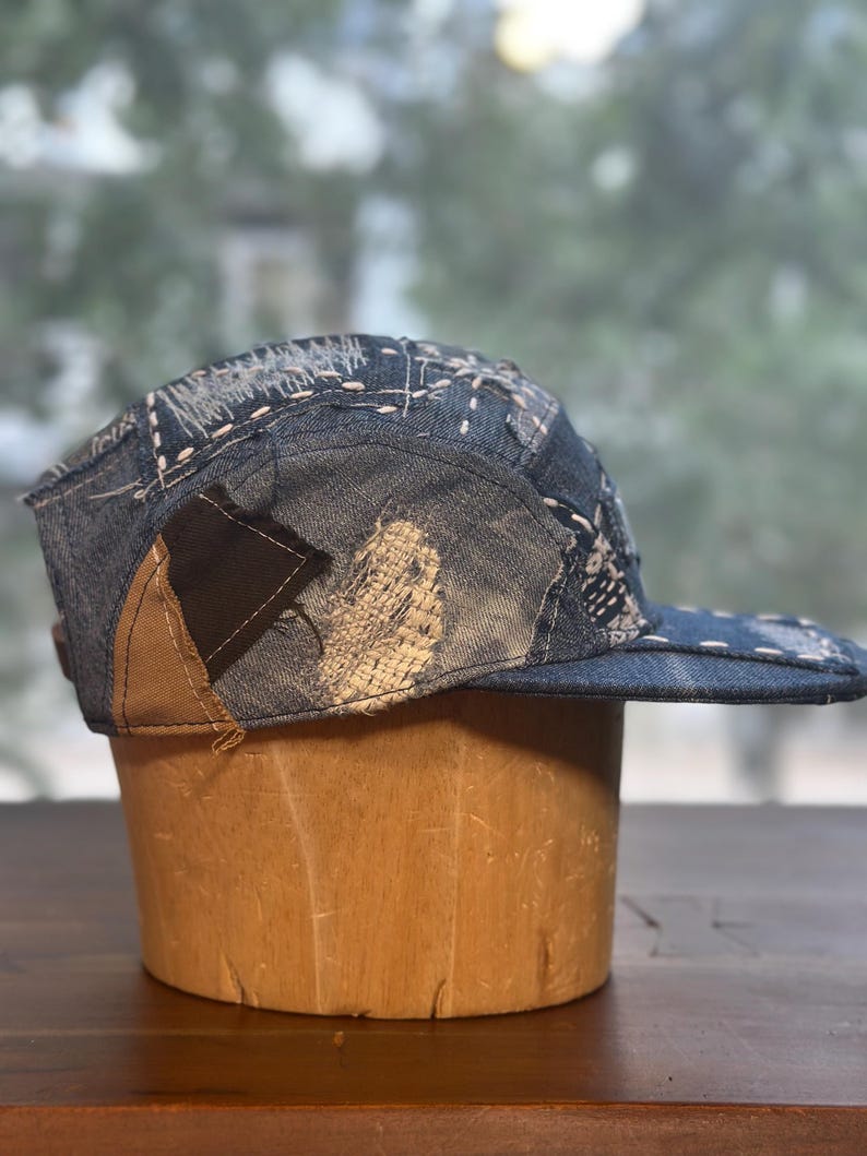 May include: A five-panel cap constructed from various shades of blue denim fabric. The hat features visible white sashiko-style stitching, frayed edges, and contrasting brown and black fabric patches integrated into the side and back panels.