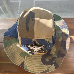 K&ouml;nnte beinhalten: Ein mehrteiliger Fischerhut aus verschiedenen Stoffresten in Camouflage, einfarbigem Beige und dunkelblauem Denim. Der Hut hat einen schwarzen gestickten Aufn&auml;her auf der Vorderseite mit dem Text CREEK RAPIDS 2020 D.O.F. TRAIL GUIDE und einem Bergsymbol.