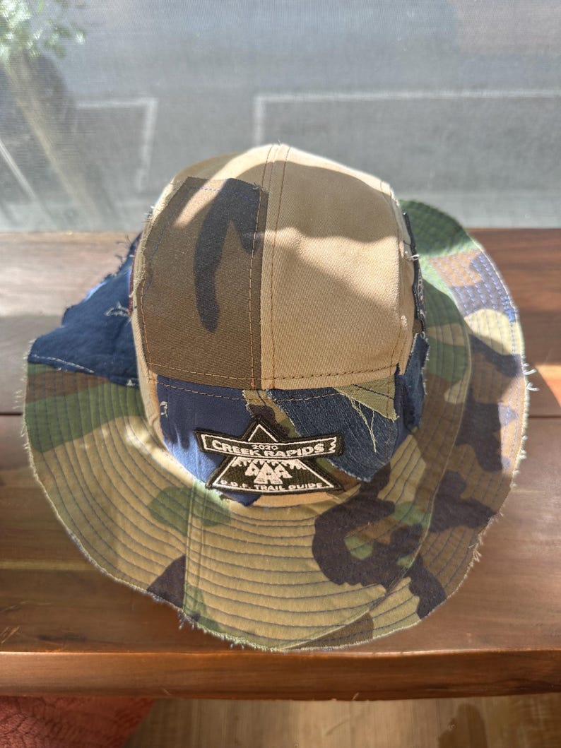 K&ouml;nnte beinhalten: Ein mehrteiliger Fischerhut aus verschiedenen Stoffresten in Camouflage, einfarbigem Beige und dunkelblauem Denim. Der Hut hat einen schwarzen gestickten Aufn&auml;her auf der Vorderseite mit dem Text CREEK RAPIDS 2020 D.O.F. TRAIL GUIDE und einem Bergsymbol.