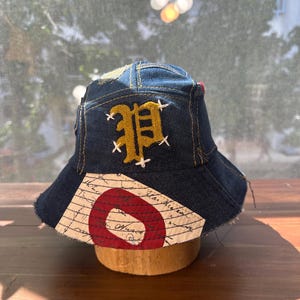May include: A dark blue denim bucket hat featuring a gold embroidered letter P with white cross-stitch detailing. The brim includes a patch of off-white fabric with black cursive script text and a large, bold red circular graphic design.