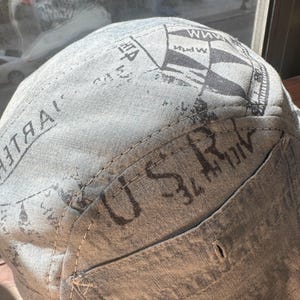 May include: A light grey fabric cap featuring a dark grey printed design with nautical flag motifs and the text U S R visible on the side panel. The cap is constructed with visible stitching and a structured crown, showcasing a textured linen-like material.