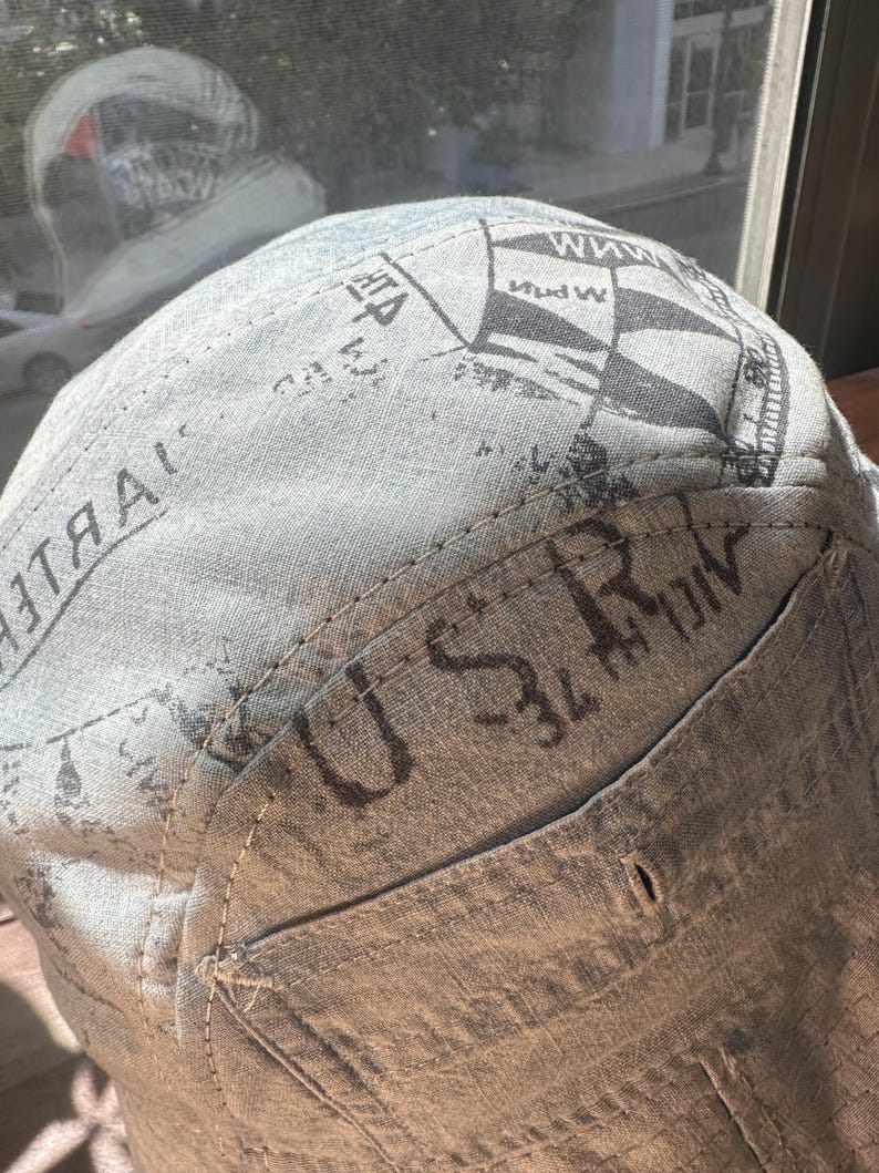 May include: A light grey fabric cap featuring a dark grey printed design with nautical flag motifs and the text U S R visible on the side panel. The cap is constructed with visible stitching and a structured crown, showcasing a textured linen-like material.