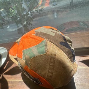 Peut inclure: Une casquette de baseball compos&eacute;e d'un assemblage de tissus vari&eacute;s, incluant du coton orange vif, de la toile beige, et des motifs camouflage vert et marron. Les panneaux sont assembl&eacute;s par des coutures visibles avec des bords effiloch&eacute;s.