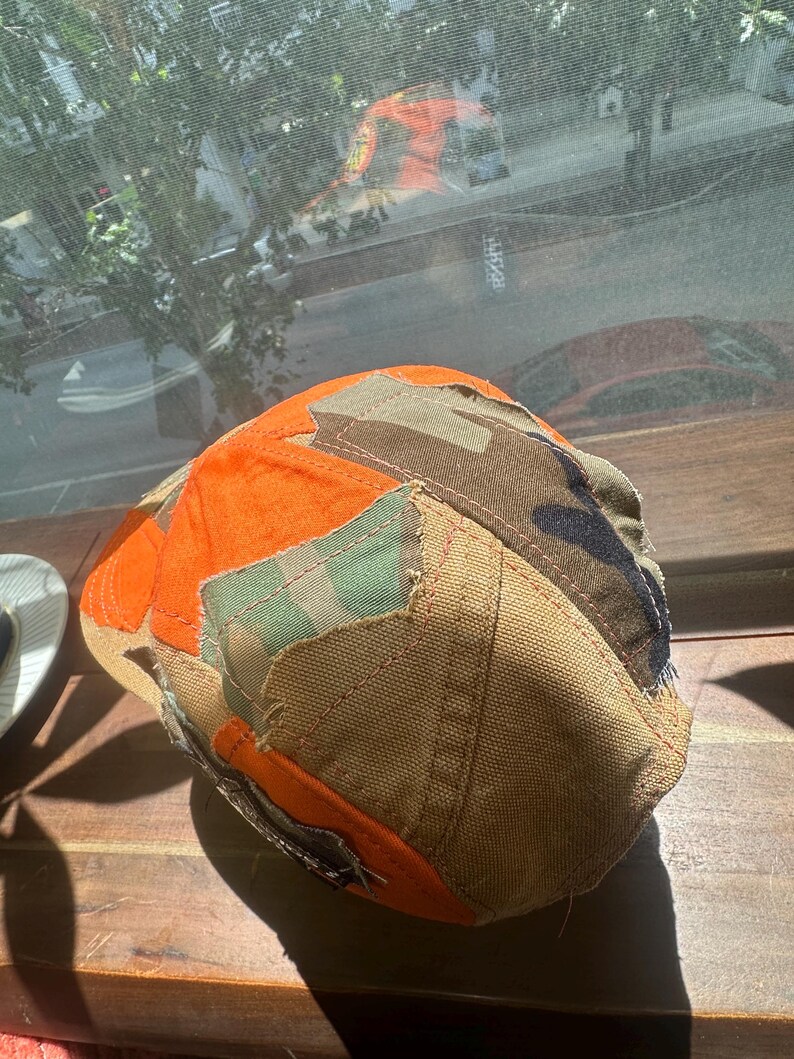 Peut inclure: Une casquette de baseball compos&eacute;e d'un assemblage de tissus vari&eacute;s, incluant du coton orange vif, de la toile beige, et des motifs camouflage vert et marron. Les panneaux sont assembl&eacute;s par des coutures visibles avec des bords effiloch&eacute;s.