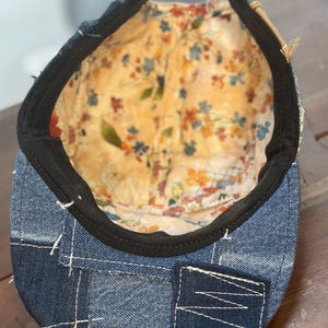 May include: A denim cap featuring a patchwork exterior with visible white stitching and a brown leather strap closure. The interior is lined with a cream-colored fabric decorated with a colorful floral pattern and finished with a black sweatband.
