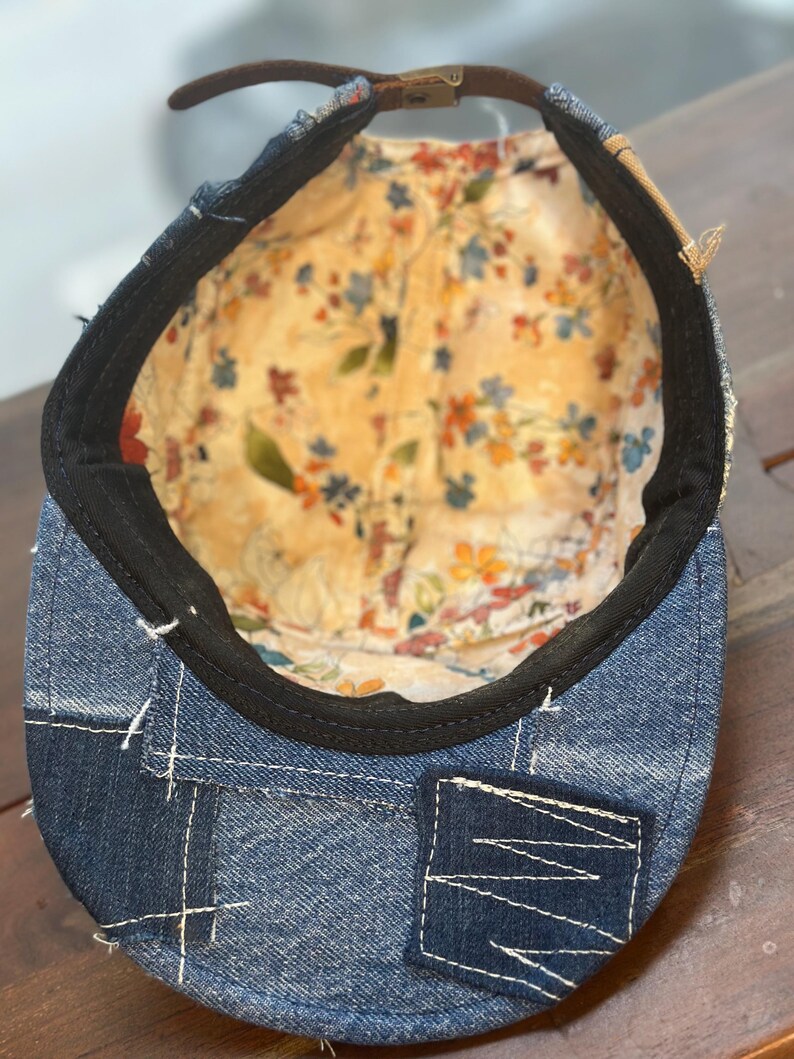 May include: A denim cap featuring a patchwork exterior with visible white stitching and a brown leather strap closure. The interior is lined with a cream-colored fabric decorated with a colorful floral pattern and finished with a black sweatband.