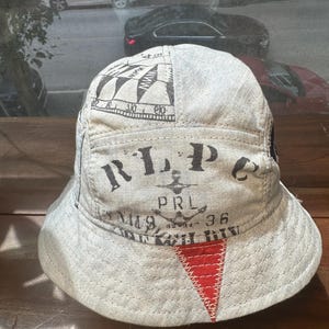 May include: A light grey bucket hat featuring black printed text that reads RLP, PRL, and 36, along with a nautical-inspired graphic. The hat includes a prominent red triangular patch stitched onto the front brim with visible white thread detailing.