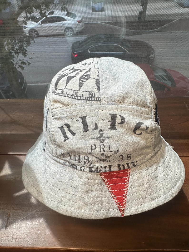 May include: A light grey bucket hat featuring black printed text that reads RLP, PRL, and 36, along with a nautical-inspired graphic. The hat includes a prominent red triangular patch stitched onto the front brim with visible white thread detailing.