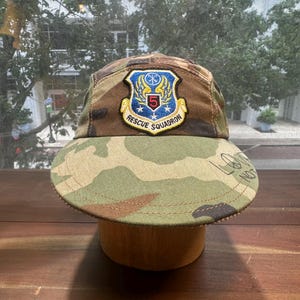 May include: A camouflage five-panel cap featuring a blue and yellow embroidered patch that reads 5 RESCUE SQUADRON. The brim displays the text LOVE NOT WAR in black lettering. The hat is constructed from a green, brown, and tan patterned fabric.