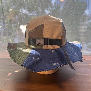 K&ouml;nnte beinhalten: Ein Patchwork-Anglerhut mit beigefarbener Krone und einer mehrfarbigen Krempe aus blauen, gr&uuml;nen und braunen Stoffbahnen. Der Hut verf&uuml;gt &uuml;ber einen schwarzen Nylonriemen mit einer Kunststoff-Steckschnalle und einen dunkelblauen Krempenbereich mit wei&szlig;en Hai-Motiven.