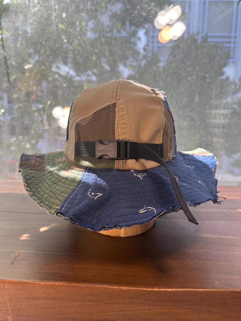K&ouml;nnte beinhalten: Ein Patchwork-Anglerhut mit beigefarbener Krone und einer mehrfarbigen Krempe aus blauen, gr&uuml;nen und braunen Stoffbahnen. Der Hut verf&uuml;gt &uuml;ber einen schwarzen Nylonriemen mit einer Kunststoff-Steckschnalle und einen dunkelblauen Krempenbereich mit wei&szlig;en Hai-Motiven.
