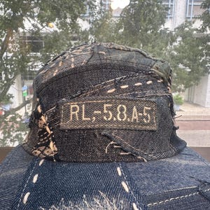 May include: A dark blue denim baseball cap featuring a patchwork design with visible light-colored stitching throughout. A rectangular fabric patch is sewn onto the front panel with the text RL,-5.8.A-5 embroidered in a light tan thread.
