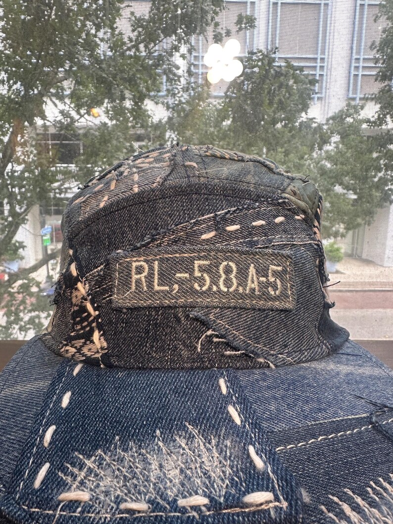 May include: A dark blue denim baseball cap featuring a patchwork design with visible light-colored stitching throughout. A rectangular fabric patch is sewn onto the front panel with the text RL,-5.8.A-5 embroidered in a light tan thread.
