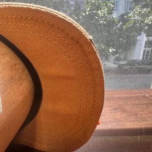 Peut inclure: Une casquette de baseball beige en tissu textur&eacute; semblable &agrave; de la toile, avec des doubles coutures visibles le long de la visi&egrave;re incurv&eacute;e. La casquette pr&eacute;sente une bande int&eacute;rieure noire et est pos&eacute;e sur un support en bois arrondi.