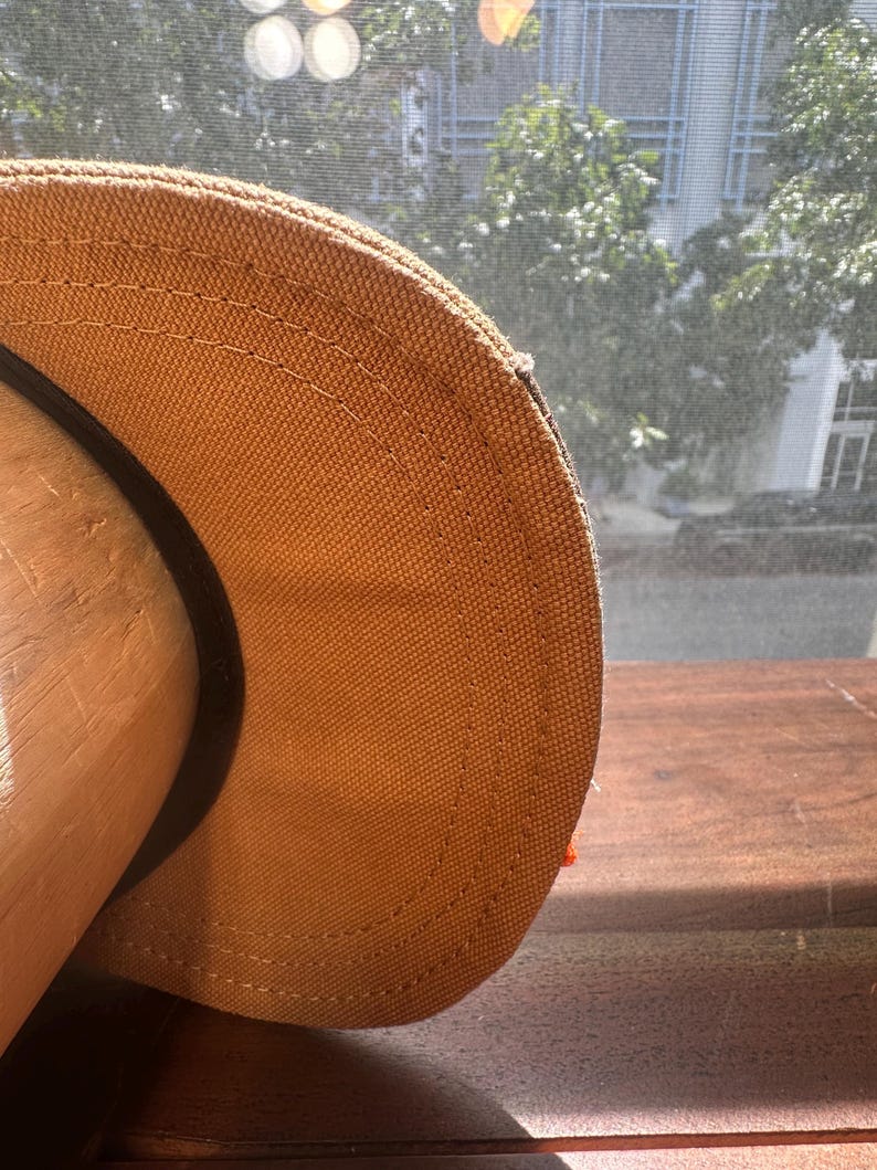 Peut inclure: Une casquette de baseball beige en tissu textur&eacute; semblable &agrave; de la toile, avec des doubles coutures visibles le long de la visi&egrave;re incurv&eacute;e. La casquette pr&eacute;sente une bande int&eacute;rieure noire et est pos&eacute;e sur un support en bois arrondi.