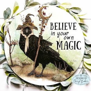 Puede incluir: Un cuervo negro con un sombrero de bruja y una bufanda, sosteniendo un bast&oacute;n con una luna creciente y hojas. El cuervo est&aacute; de pie sobre un fondo marr&oacute;n y verde con el texto "BELIEVE in your own MAGIC" y la firma del artista.