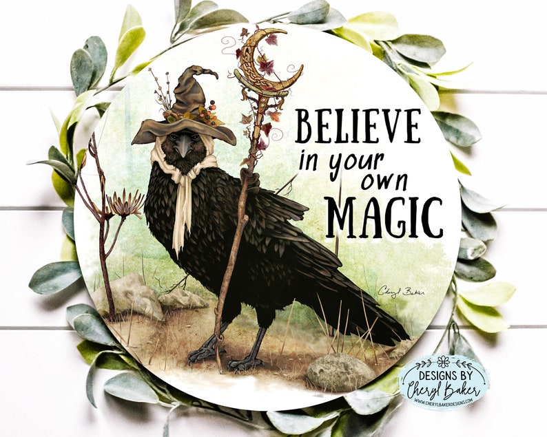 Puede incluir: Un cuervo negro con un sombrero de bruja y una bufanda, sosteniendo un bast&oacute;n con una luna creciente y hojas. El cuervo est&aacute; de pie sobre un fondo marr&oacute;n y verde con el texto "BELIEVE in your own MAGIC" y la firma del artista.