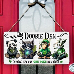 May include: A white wooden sign with the text "The Doobie Den" and a cartoon image of a panda, raccoon, monkey, and rabbit smoking. The sign also says "Sorting life out one toke at a time".