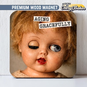 May include: Square wood magnet with a vintage doll's face, one eye open and the other closed, and red lips. The text "AGING GRACEFULLY" is displayed across the doll's face. The magnet is labelled "PREMIUM WOOD MAGNET" and "DESIGNS BY Cheryl Baker".
