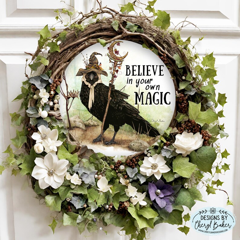 Puede incluir: Una corona hecha de enredaderas y vegetaci&oacute;n con flores blancas y bayas marrones. Un letrero redondo con un cuervo negro que lleva un sombrero de bruja y sostiene un bast&oacute;n est&aacute; en el centro de la corona. El letrero dice "Cree en tu propia magia".