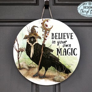 Puede incluir: Un letrero redondo de madera con un cuervo negro que lleva un sombrero de bruja y sostiene un bast&oacute;n. El letrero tiene un fondo verde y marr&oacute;n con el texto "Cree en tu propia magia" en letras negras.